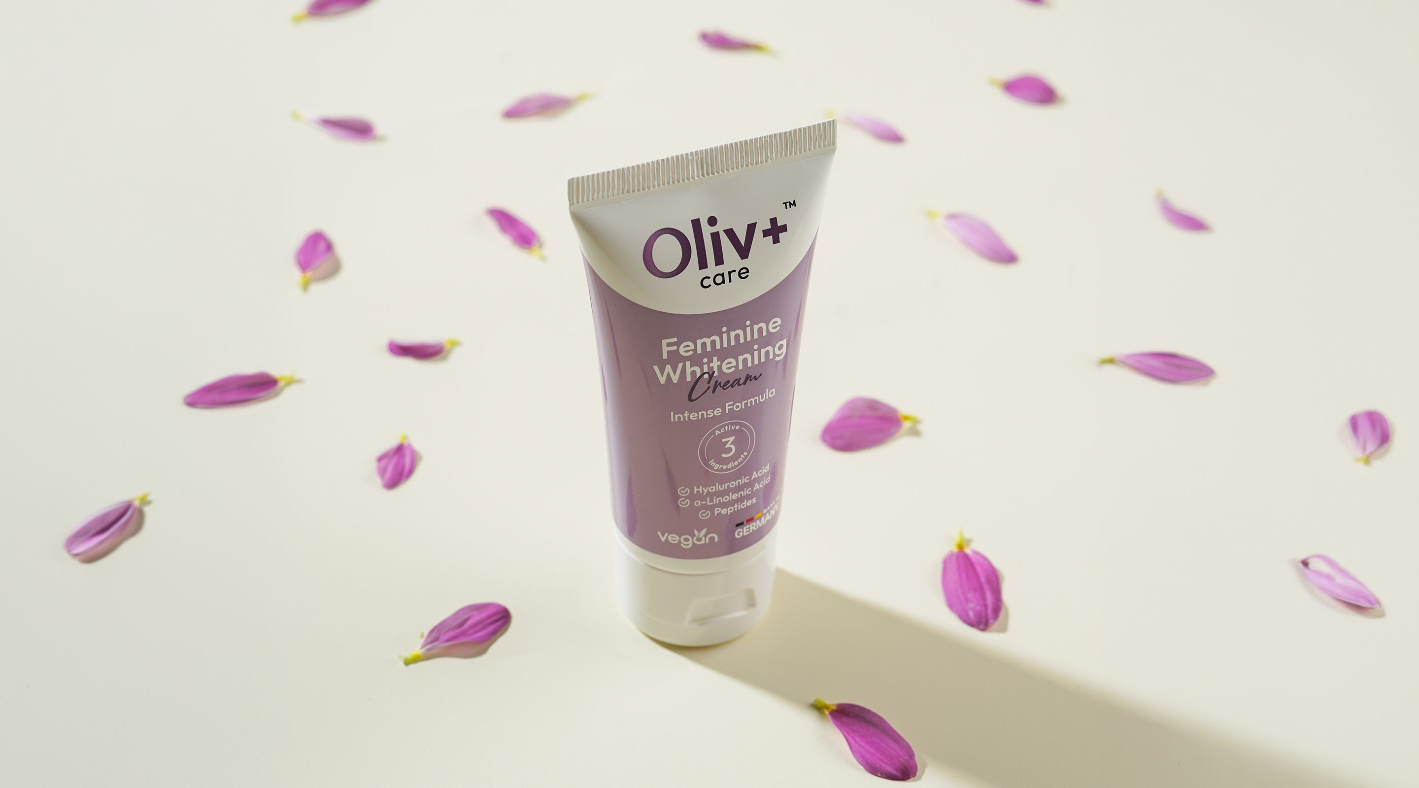 Kemasan Olivplus care Feminine Whitening Cream di atas latar krem dengan kelopak bunga ungu, menunjukkan produk vegan yang aman untuk sehari-hari.