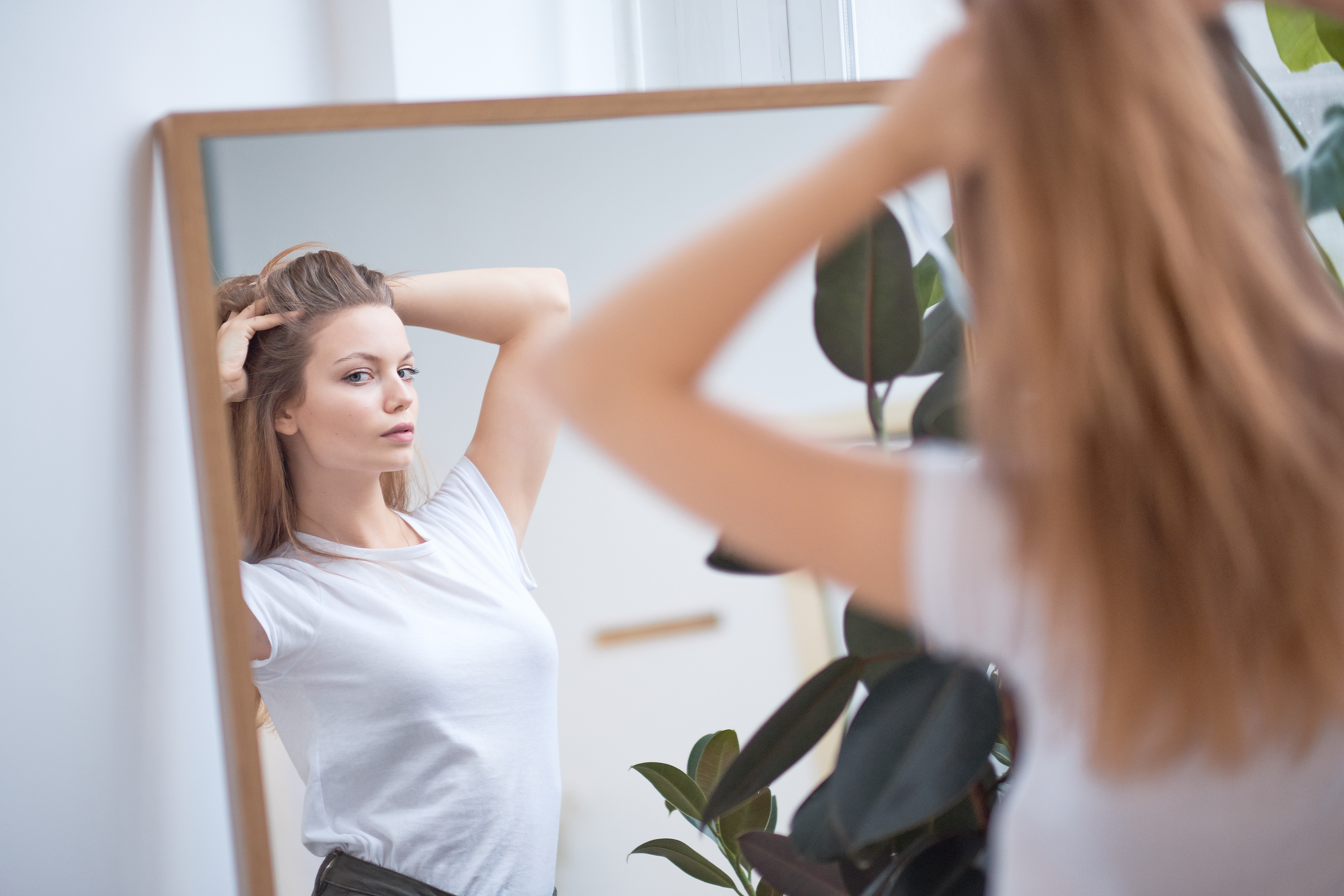 A young woman in a white t-shirt checking her hair in a mirror with a neutral expression.